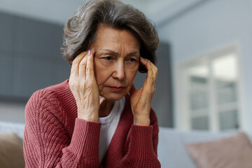 Unhappy senior woman touching temples, feeling strong headache or high blood pressure, sitting on...