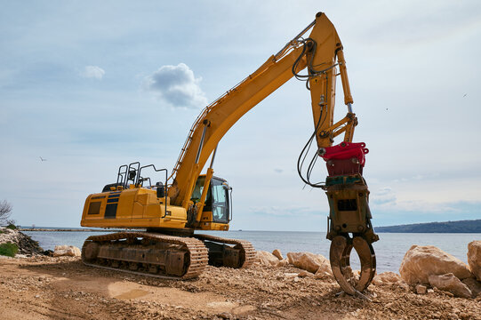 Yellow tracked excavator with hydraulic claw attachment working on a rocky coastal construction site near the sea under a partly cloudy sky