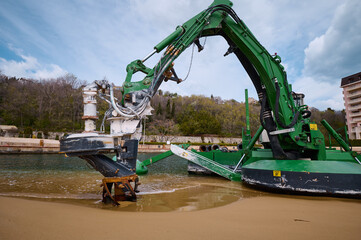 Green amphibious dredging machine working at the edge of a beach and water channel, used for underwater excavation and maintenance near a marina or coastal area