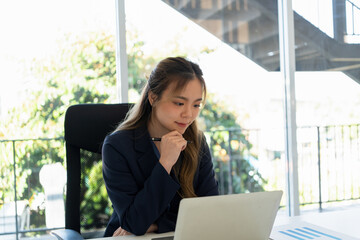 Fototapeta premium Businesswoman sitting at desk on couch in workplace or at home working on laptop and analyzing data on charts and graphs and writing on papers to make business plan and strategies for company.