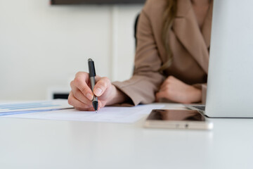 Businesswoman sitting at desk on couch in workplace or at home working on laptop and analyzing data on charts and graphs and writing on papers to make business plan and strategies for company.