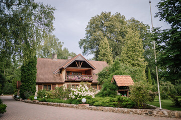 Charming countryside house with tiled roof, wooden balcony, blooming flowers, lush garden, and stone-paved driveway surrounded by tall trees..