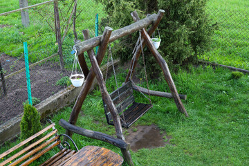 A handmade wooden swing stands on a green lawn. The swing has a bench made of dark planks, suspended by metal chains from roughly processed wooden supports.