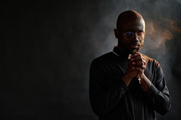 African American priest in black cassock holding rosary with golden cross in hands while praying silently in room with incense smoke in air
