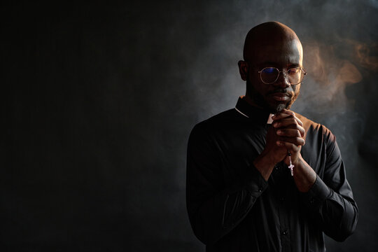 African American priest in black cassock holding rosary with golden cross in hands while praying silently in room with incense smoke in air - Powered by Adobe