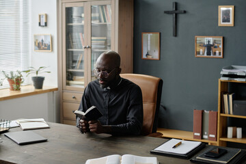 Young African American priest in black cassock sitting at wooden table and reading bible. Office...
