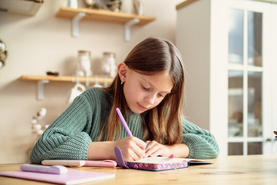 A Young Girl Writes In Her Journal At The Table During The Day Inside The Kitchen