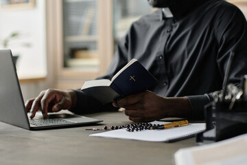 Medium close up of unrecognizable pastor in black cassock sitting at wooden desk typing on keyboard and holding bible while working in office