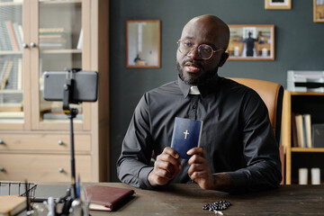 Medium shot of African American pastor in black cassock holding small bible with cross on cover...