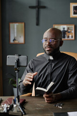 Vertical shot of African American priest looking at camera of smartphone while reciting bible...