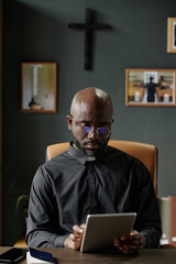 Vertical shot of African American pastor using tablet to check email while sitting at table in...