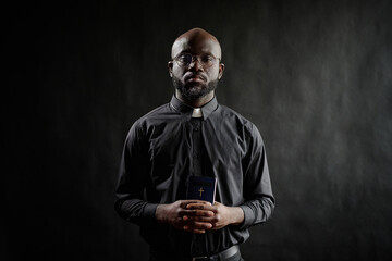 Portrait of solemn young adult pastor wearing black cassock, holding small bible while posing for photo with jet black background