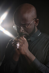 Vertical shot of young African American pastor with folded hands in prayer holding black rosary with silver cross standing stull while praying silently
