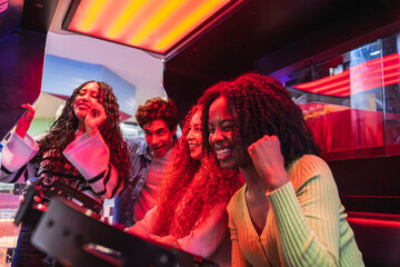 Group of young friends playing and having fun together with arcade video game inside a bowling alley, illuminated by colorful neon lights