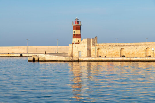 Red and white lighthouse (Faro Rosso) on the ancient pier in the city of Monopoli, Bari, Italy during last hours of sun of the day