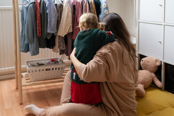 Authentic cozy moment of a mother and her little daughter hugging at home, surrounded by children's clothes and toys. Real life, real emotions, real love.