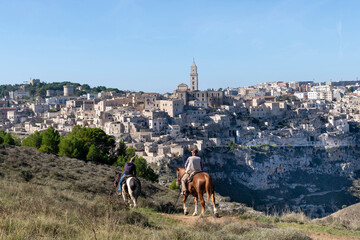 Two man and horses riding along ridge with panoramic view of cliffs with buildings on the Gravina di Matera of Matera, Italy, know for the Sassi or Ancient town