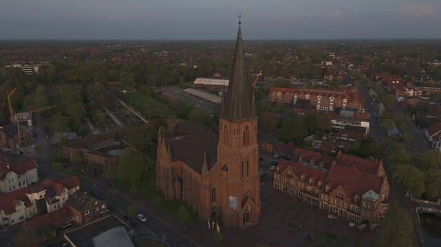 Orbiting drone shot of the red-brick St. Antonius Church in Papenburg, Germany, during golden hour. Surrounded by rooftops, trees and evening light. Historic architecture in a peaceful townscape.