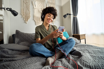 Young happy funky African American woman, gen z Afro girl wearing headphones using smartphone at home sitting on bed looking at mobile phone listening music in apps online on cellphone in bedroom.