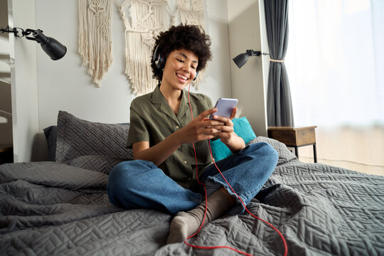Young happy funky African American woman, gen z Afro girl wearing headphones using smartphone at home sitting on bed looking at mobile phone listening music in apps online on cellphone in bedroom.