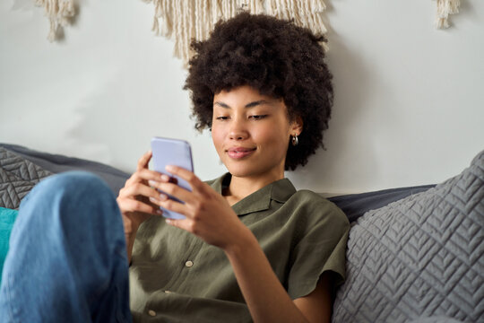 Young relaxed happy African American woman, pretty female gen z student using smartphone at home sitting on bed looking at mobile phone buying online in shopping apps on cellphone in bedroom.