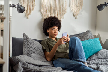 Young African American woman, pretty generation z girl student using smartphone at home sitting on bed looking at mobile phone scrolling social media apps online on cellphone relaxing in bedroom.