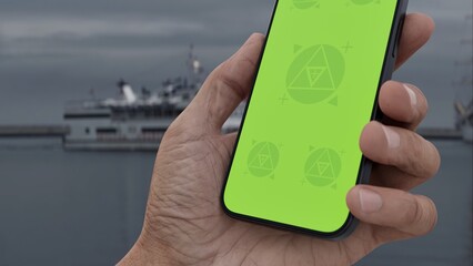 GREEN SCREEN CHROMA KEY Hand holding smartphone with green screen at a harbor on an overcast day,...