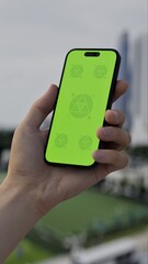 GREEN SCREEN CHROMA KEY Person holding a smartphone with a green screen outdoors, overlooking a modern urban skyline and grassy area on a cloudy day