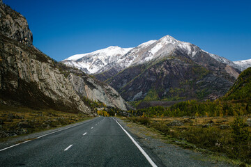 A mountain road winding through the rugged terrain of the Altai Republic, surrounded by harsh, snow-covered peaks and dramatic natural beauty.