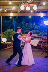 Bride and groom share a romantic first dance under glowing lights in an outdoor venue, surrounded by vintage d&eacute;cor and warm evening ambiance..