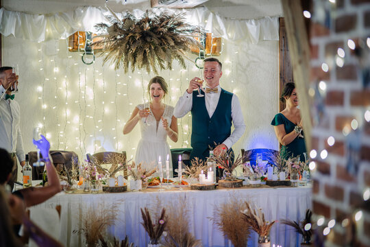 Bride and groom share a joyful toast at the wedding head table, surrounded by fairy lights, rustic decor, and smiling guests during a festive reception..