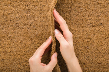 Young adult woman hands holding and showing thickness orthopedic mattress brown coconut coir. Closeup. Point of view shot. Top down view.