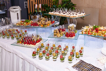 Elegant buffet table with assorted fruit skewers, canapes, mini appetizers, and champagne bottles arranged on glass stands for a formal celebration or corporate event