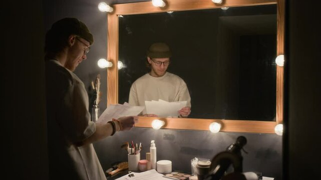 Young man fixing his beanie while looking at mirror and rehearsing his stand-up script in dimly lit dressing room