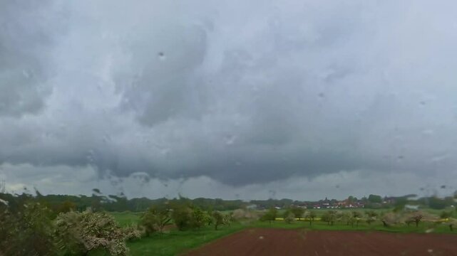 Nahaufnahme von Regentropfen, die an einer Fensterscheibe runterlaufen, mit dramatisch dunklen Regenwolken &uuml;ber Feldern und Dorf am Horizont, tr&uuml;be Wetterstimmung,  Metrologie, Melancholie, Herbst