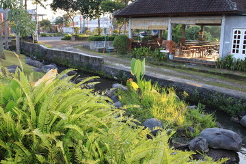 Lush green garden with a small stream running through it. The garden is surrounded by a stone wall and has a wooden pavilion in the background