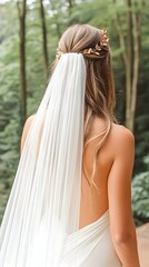 Back view of bride in flowing white dress and veil, adorned with gold hairpiece, standing in a forest