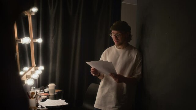 Young man holding his stand-up scrip and rehearsing lines out lout, leaning on wall of dimly lit dressing room before going on stage