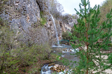 A picturesque waterfall flowing from a rocky cliff into a mountain river surrounded by forest in spring.