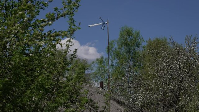 Outdoor passive television decimeter antenna on a long pole among trees against a blue sky near a one-story private house with a slate roof. Analog and digital radio signal receiver in remote areas.