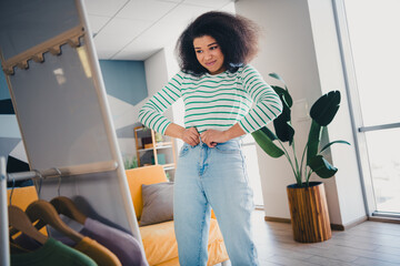 Young woman adjusting her jeans in bright and stylish living room with cozy furniture and green...