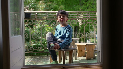 Young boy sitting on a wooden stool on a balcony, smiling and holding his foot, surrounded by...