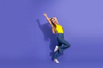 Fotobehang Young woman in bright yellow clothing and sunglasses posing playfully against a vibrant purple background © deagreez