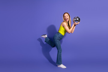 Joyful young woman posing playfully with a disco ball on a vibrant purple background in stylish...