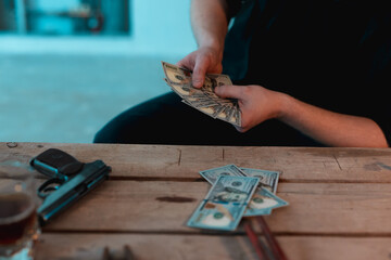 Cropped photo of man in a black clothes counting money in the basement. Gun and glass with alcohol...