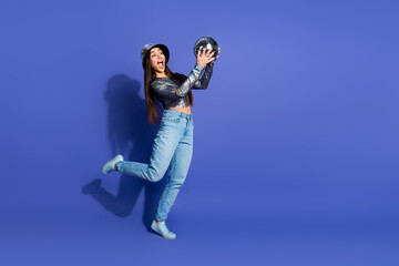 Obraz premium Young woman posing with a fun attitude holding a shiny disco ball against a vibrant purple background wearing trendy casual fashion.