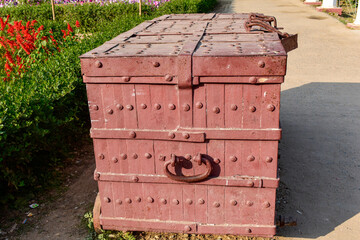 The image features a vintage iron chest along a garden path, surrounded by flowers. Its rustic maroon colour and sturdy handles suggest stories of forgotten treasures, blending history with nature. 