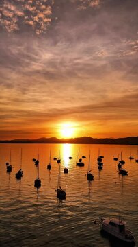 Vertical video of drone flying over beautiful Lake Garda in Italy, colorful red orange  gold sunset, sun reflection on water, many boats on water, mountains visible on horizon, blue sky