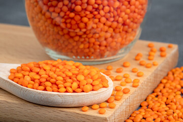 On a chopping board, there is a spoon filled with orange lentils and a glass jar full of red lentils
