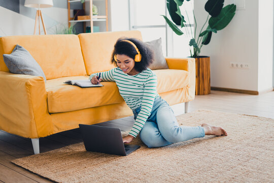 Young woman in cozy living room enjoying leisure time with laptop, headphones, and a book, sitting comfortably on rug near couch.